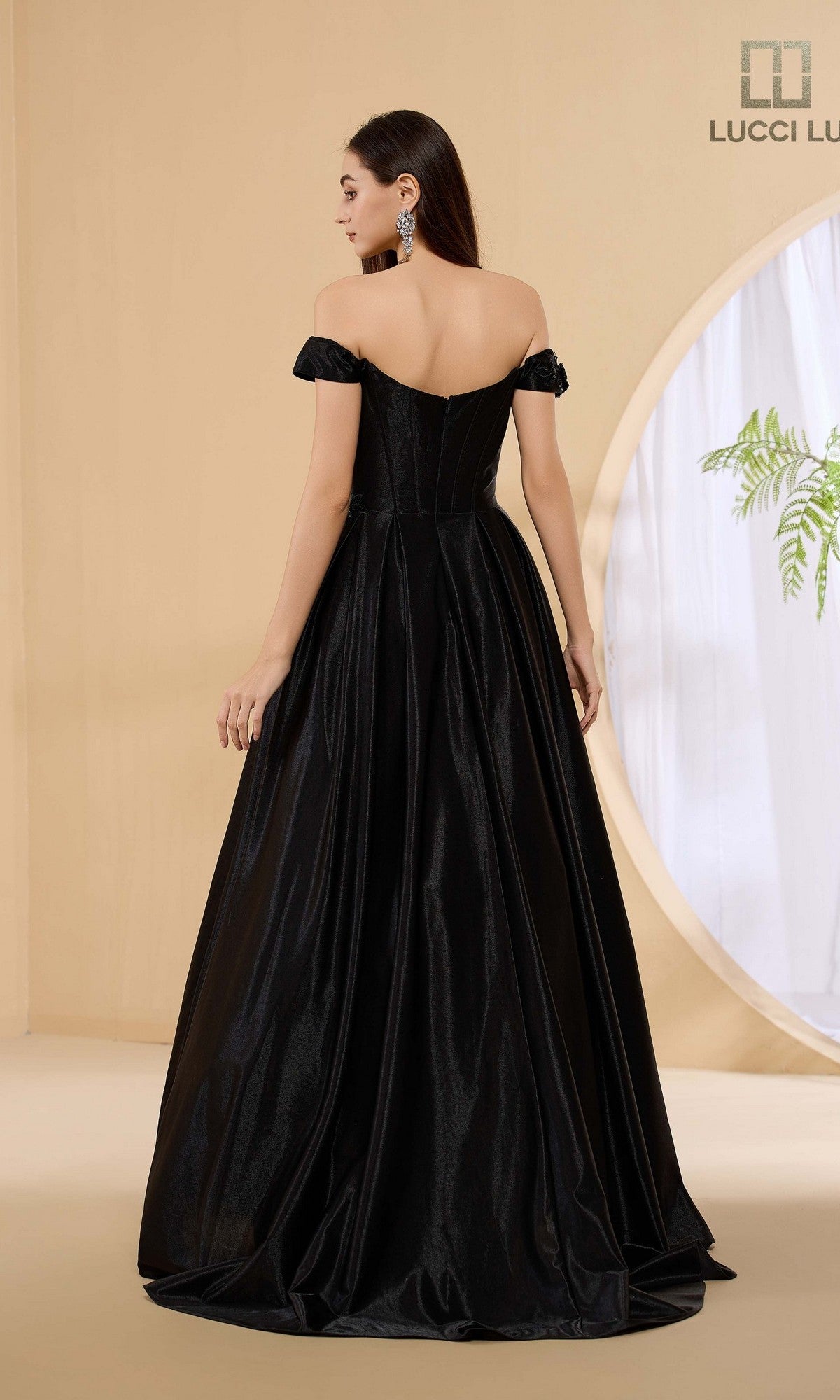 A woman faces away from the camera in a minimalist room, modeling the Lucci Lu 1472 long off-the-shoulder A-line prom dress, with beige walls and a round window in the background.